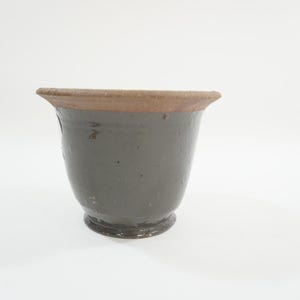 May include: A dark gray ceramic planter with a flared rim and a footed base. The rim has a brown band. The planter has a slightly textured surface and is suitable for indoor or outdoor use. A decorative item for plants.