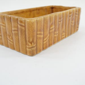 May include: A rectangular, ceramic planter with a bamboo-style design. The planter is a warm, light brown color and has a textured surface. The interior is empty, and the planter sits on a white surface.