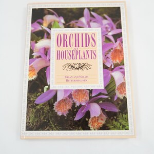 May include: A hardcover book titled "Orchids as Houseplants" with a floral cover design. The cover features a close-up of vibrant purple and orange orchids. The book has a cream-colored border and a white background.