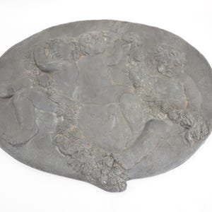 以下が含まれることがあります： グレーの楕円形のレリーフ彫刻で、古典的なスタイルで複数の人物、おそらくケルビムが描かれています。人物はダイナミックな構図で表現され、詳細なテクスチャと形状が特徴です。彫刻はおそらく石または同様の素材で作られています。