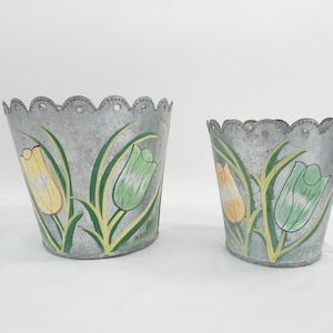 May include: Two galvanized metal planters with a scalloped top edge. Each planter features a hand-painted design of yellow and green tulips with long green leaves. The metal has a mottled gray finish, and the containers are tapered in shape.