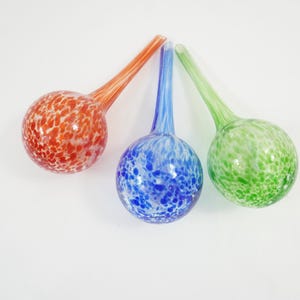Puede incluir: Tres globos de riego de vidrio con cuellos largos y estrechos y bases esféricas. Cada globo presenta un patrón moteado distintivo en naranja, azul o verde, con acentos blancos en todo el vidrio. Los globos están dispuestos en forma de abanico.