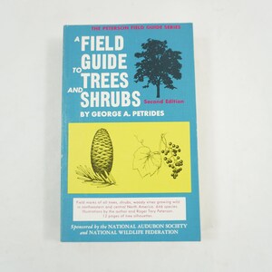 Könnte beinhalten: Ein blauer Buchumschlag mit dem Titel "Field Guide to Trees and Shrubs" von George A. Petrides. Der Umschlag zeigt Illustrationen einer Kiefernzapfen, eines Baumes und einer Rebe mit Beeren.
