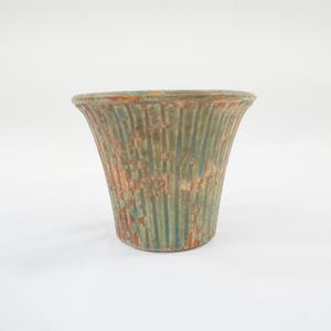 May include: A flared ceramic planter featuring a vertical ribbed texture. The exterior surface displays a weathered finish with a blend of muted sage green and earthy terracotta orange tones, creating a mottled appearance across the entire vessel.
