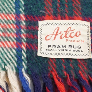 May include: Close-up of a vintage Artco Products pram rug. The rug features a classic plaid pattern in red, green, white, and blue. The label reads "PRAM RUG 100% VIRGIN WOOL". The rug has a fringed edge.