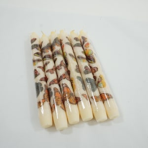 May include: A collection of seven cream-colored taper candles. Each candle is decorated with a repeating pattern of orange, brown, and gray butterflies. The candles are arranged diagonally against a white background.