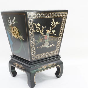 May include: A black lacquered planter with a tapered square shape, featuring inlaid mother-of-pearl floral motifs and a figure standing on a bridge. The piece rests on a four-legged base with gold-painted accents and includes a circular brass handle.