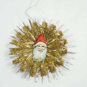 Op de afbeelding: Een vintage kerstversiering met een kerstmangezicht in het midden, omringd door gouden en zilveren slingers. De versiering heeft een sterrenburst-ontwerp en een zilveren touwtje om op te hangen. Een feestelijke vakantieversiering.