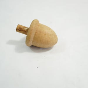 May include: A wooden acorn-shaped object with a cylindrical stem. The acorn cap is rounded and the stem is a lighter shade of brown. The object is made of light-colored wood and sits on a white surface.