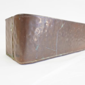 May include: A rectangular copper container with a hammered texture and visible patina. The object features a vertical seam along the rounded corner and a metallic brown finish with subtle green oxidation marks across the surface.