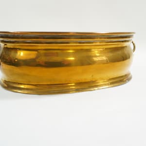May include: A vintage, oval-shaped brass planter with two small handles. The planter has a shiny, golden finish and a slightly curved base. It is likely used for displaying plants or flowers.