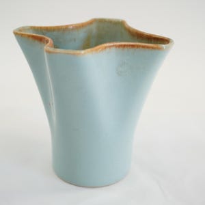 May include: A light blue ceramic vase with a unique, wavy rim and a flared shape. The rim has a brown trim. The vase has a smooth, matte finish and is suitable for displaying flowers or as a decorative accent piece.