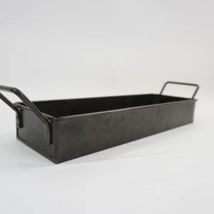 May include: A rectangular black metal tray with a smooth finish and two angled handles attached to the short sides. The tray features a deep, open basin design with straight walls and a flat base, constructed from dark, solid metal material.