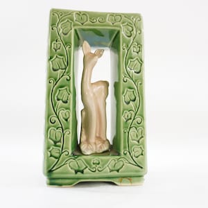 May include: A green ceramic wall pocket with a rectangular frame and a deer figurine inside. The frame is decorated with a leaf and vine pattern. The deer is light brown and stands inside the frame.