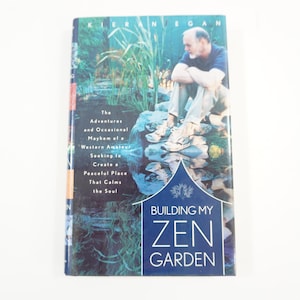 Pode incluir: Uma capa de livro com fundo azul e o título "Building My Zen Garden". A capa apresenta uma foto de um homem sentado em rochas perto de um riacho em um jardim verde exuberante.