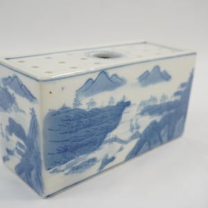 May include: A rectangular ceramic flower frog with a white base and blue hand-painted landscape scenes featuring mountains, trees, and water. The top surface is flat with a series of small circular holes for arranging stems in a grid pattern.