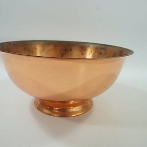 May include: A polished copper bowl with a wide, open top and a rounded base. The exterior surface features a warm, metallic orange-brown tone, while the interior shows signs of natural oxidation and dark spots along the inner rim and bottom.