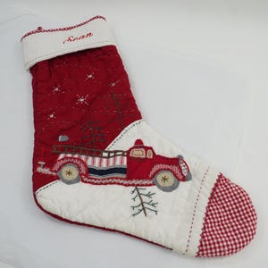 Op de afbeelding: Een gewatteerde kerstsok in rood en wit met een geborduurde rode brandweerwagen die een boom vervoert. De witte boord is geborduurd met de naam Sean in rood schrift. Het teengedeelte heeft een rood-wit ruitpatroon in vichy-stijl.