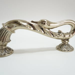 Può includere: Maniglia per armadio color argento con design a cigno. La maniglia si curva con grazia, con una testa di cigno e dettagli di foglie. La base ha un design floreale ornato. Un elemento decorativo per l'hardware.