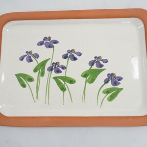 May include: Rectangular ceramic tray with a terracotta-colored border. The tray's interior is white, adorned with a floral design of purple irises and green leaves. A decorative piece for serving or display.