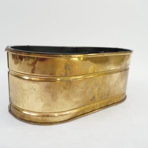Puede incluir: Una maceta ovalada de latón con un exterior dorado pulido y reflectante que presenta una sutil textura martillada. El recipiente tiene un borde liso y redondeado y un interior oscuro y profundo, mostrando una construcción metálica.