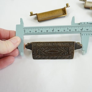 May include: A close-up of a decorative brass drawer pull with an ornate design. The pull is being measured with a ruler, showing it is approximately 3.5 inches long.