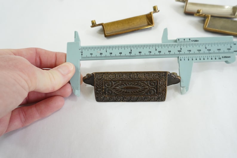 May include: A close-up of a decorative brass drawer pull with an ornate design. The pull is being measured with a ruler, showing it is approximately 3.5 inches long.