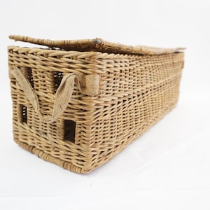 May include: A rectangular woven wicker basket featuring a hinged lid and two light tan fabric handles attached to the front side. The basket has a natural light brown finish with a visible woven texture and small square openings on the front.
