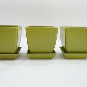 Puede incluir: Tres macetas cuadradas de cerámica verde oliva con platillos cuadrados a juego. Las macetas tienen una superficie texturizada y un diseño ligeramente cónico. Ideales para plantas de interior o exterior.
