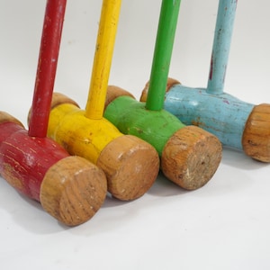 May include: Four colorful croquet mallets with wooden heads and handles. The mallets are painted red, yellow, green, and blue, with visible wear and tear, suggesting they are vintage or well-used. The wooden heads are a natural wood color.