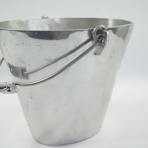 Puede incluir: Un cubo de metal plateado con un asa curva. El cubo tiene una forma cónica, más ancho en la parte superior y más estrecho en la base. El asa está sujeta con presillas metálicas.