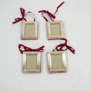 Puede incluir: Cuatro marcos de fotos rectangulares pequeños de tono plateado dispuestos en cuadrícula. Cada marco presenta un diseño de borde escalonado y una cinta roja oscura atada en un lazo en la parte superior para colgar. El centro tiene un inserto beige.