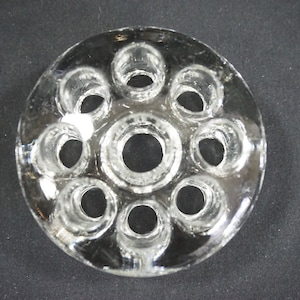 May include: A clear glass flower frog with 10 round holes for holding stems. The frog is circular and has a smooth, polished surface.