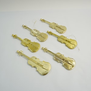 May include: Six gold-colored violin ornaments are arranged on a white surface. Each ornament has a detailed design, including the strings and the shape of the instrument. The ornaments are likely for decoration.