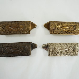 May include: Four ornate brass drawer pulls with a decorative floral design. The pulls are rectangular in shape and have a slightly raised centre section. Two of the pulls are a darker brass colour, while the other two are a lighter brass colour.