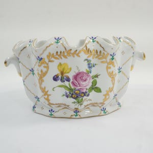 May include: White ceramic bowl with a scalloped rim and two handles. The bowl is decorated with gold trim, a floral design featuring a pink rose, yellow iris, and blue flowers. The bowl has a diamond pattern.