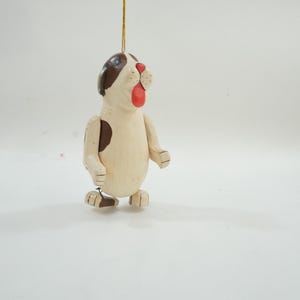 May include: Wooden dog ornament with a brown and white color scheme. The dog has a brown spot on its side and head, with a red mouth. The ornament is suspended by a gold string, and is standing on its feet.
