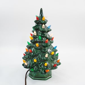 May include: A dark green ceramic Christmas tree lamp with a glossy finish, featuring a tiered branch design and a hexagonal base. The tree is decorated with numerous small, translucent plastic bulbs in red, yellow, blue, and clear colors.