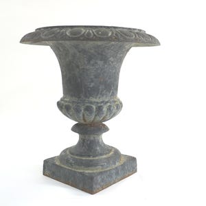 Könnte beinhalten: Eine verwitterte, graue Gusseisenurne mit einem breiten, dekorativen Rand und einem Sockel. Die Urne hat ein klassisches, antikes Design, geeignet für die Außendekoration im Garten. Der Sockel ist quadratisch und die Urne ist etwa 46 cm hoch.