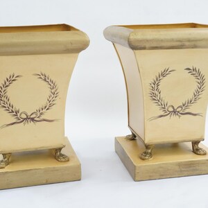 May include: A pair of cream-coloured square planters with a flared top rim and a base supported by four small claw feet. Each planter features a dark brown painted laurel wreath design with a bow on the front panel, finished with a distressed gold trim.