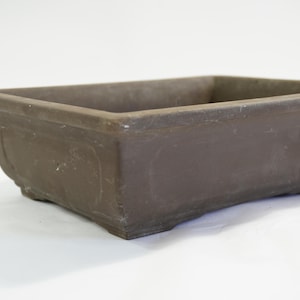 以下が含まれることがあります： わずかにテクスチャ加工された表面を持つ、長方形の茶色の盆栽鉢。 鉢は、隆起した縁と緩やかに湾曲したベースを備えた、シンプルでクラシックなデザインです。 盆栽やその他の小さな植物に最適です。