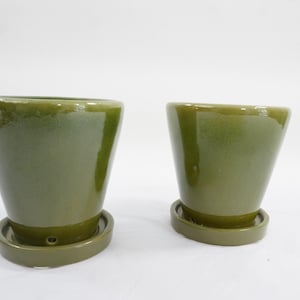 May include: Two olive green ceramic planters with matching saucers. The planters have a glossy finish and a slightly flared shape. The saucers are round and have a lip to catch excess water. Ideal for indoor or outdoor use.