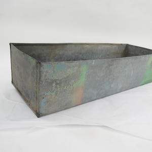 Puede incluir: Una jardinera rectangular de metal galvanizado con aspecto envejecido. La caja tiene un color base gris con toques de pintura azul, verde y naranja en el lateral. La jardinera está vacía y se encuentra sobre una superficie blanca.