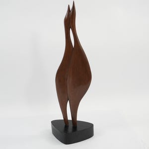 May include: A tall, smooth, dark brown wooden sculpture depicting two elongated, abstract bird-like figures standing side-by-side. The figures are joined at the upper necks and rest upon a solid, triangular black base with rounded edges.