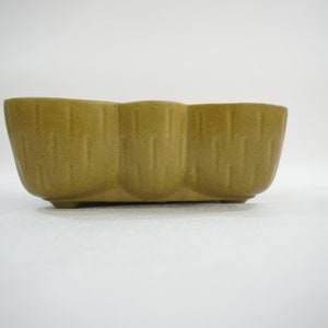May include: A green ceramic planter with a unique, textured design. The planter has three rounded sections and a rectangular base.