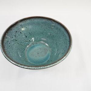 May include: A round ceramic bowl featuring a speckled teal and blue glaze with a dark brown rim. The interior surface displays a mottled texture with varying shades of blue, transitioning from a deep teal at the edges to a lighter blue center.