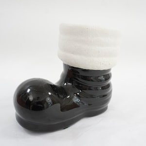May include: A ceramic boot-shaped planter, primarily black with a glossy finish. The top of the boot features a textured, white cuff. The planter is designed to hold small plants or flowers, adding a whimsical touch to home decor.