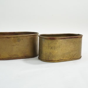 May include: Two antique brass planters with an oval shape. The planters have a weathered, aged appearance with a warm, golden-brown color. The edges are slightly darker, adding depth to the design. These planters are suitable for indoor or outdoor use.