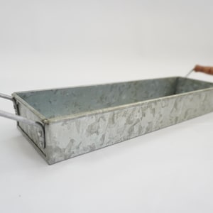 May include: A rectangular galvanized metal tray with a mottled silver finish. The tray features two metal handles on opposite ends, each equipped with a cylindrical brown wooden grip for handling. The structure is open-topped and shallow.