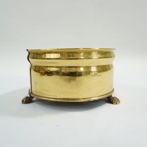 Può includere: Un piccolo contenitore rotondo in ottone con finitura lucida. Il contenitore poggia su tre piccoli piedini decorativi. Il bordo del contenitore ha un piccolo labbro. Probabilmente è un oggetto antico o vintage.